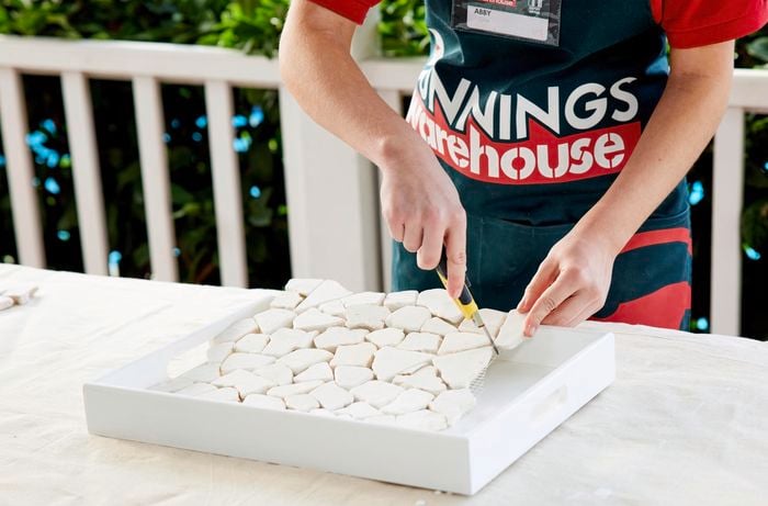 A woman places mosaic tiles onto a tray and cuts off extra tiles.