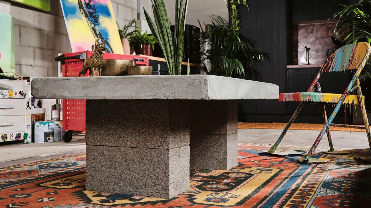 A concrete table with a plant, an ornate rug, and a colourful, paint-splattered chair in an artistic setting.