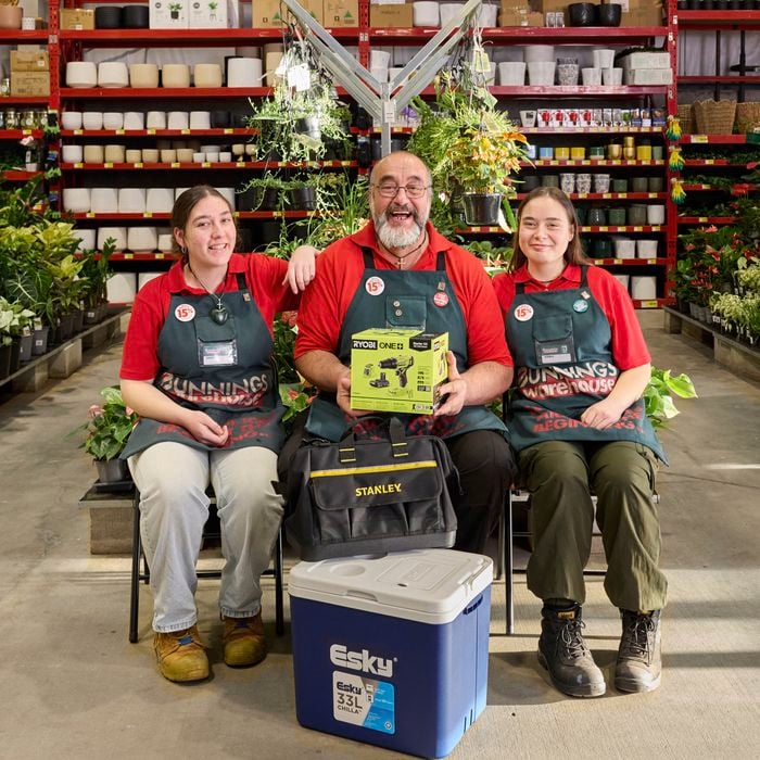 Three Bunnings team members seated in a store aisle lined with plants and pots. The person in the centre holds a Ryobi ONE+ tool kit, with a Stanley tool bag and a blue Esky cooler positioned in front. All three are wearing red shirts and Bunnings aprons, with store shelving and hanging plants visible in the background.