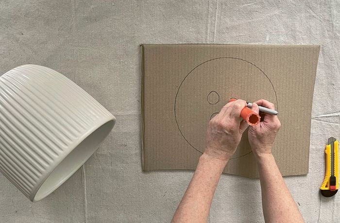 Hands using a marker to trace around an orange conduit offcut onto cardboard, next to a white pot and a utility knife.