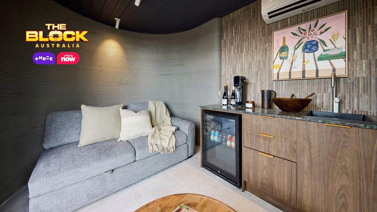 A room with a couch, bar fridge, coffee table, a picture split system on the wall in it.