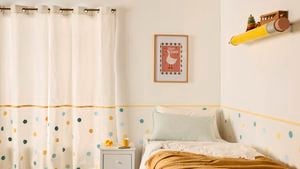 A large yellow pencil shelf topped with wooden toys on a neutral wall with colourful polka dots with a bed with pillows and a cover on it, in the bedroom.