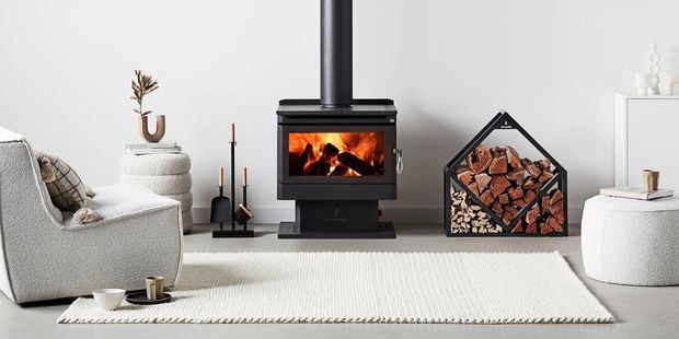 A room with an indoor wood heater, pieces of wood, wood heater accessories, a rug and a chair in it. 