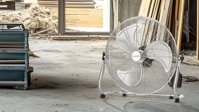 Large silver floor fan positioned inside a construction site, with stacks of materials, exposed walls, and building work visible in the background.