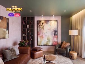A modern living room featuring a large abstract painting on the wall, a brown leather sofa with cushions, an armchair, a wooden side table, and a floor lamp. A bookshelf with books and decorative items stands nearby. The room has soft lighting and curtains covering the windows. 