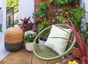 A balcony with potted plants, a wicker stool, and a green chair with a striped cushion, against a wooden screen with hanging plants.