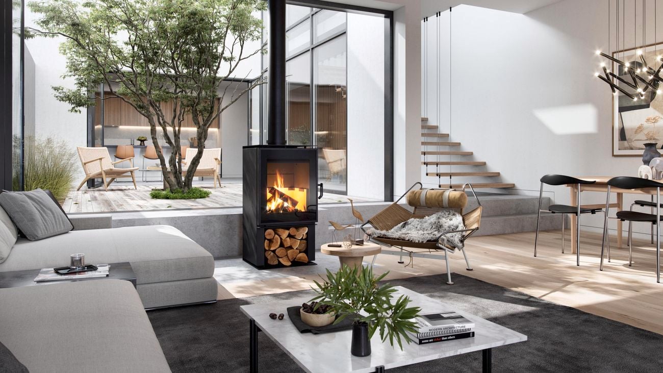 Wide shot of a living room with an indoor fire pit at the centre, with a grey couch,rug and glass coffee table in the foreground.