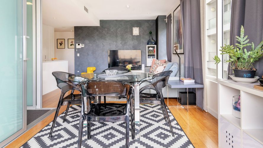 Contemporary open-plan dining and living space featuring a glass dining table, transparent chairs, patterned rug, large windows and a lounge area with a television and grey accent wall.