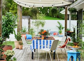 Outdoor dining area on a timber deck with a blue and white striped tablecloth, surrounded by potted plants and chairs, with a shade sail overhead