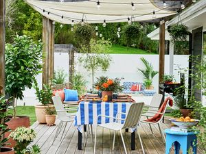 Outdoor dining area on a timber deck with a blue and white striped tablecloth, surrounded by potted plants and chairs, with a shade sail overhead