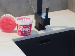 A pink Scrub Mommy sponge and a tub of The Pink Stuff cleaning paste sitting on a benchtop beside a black tap.