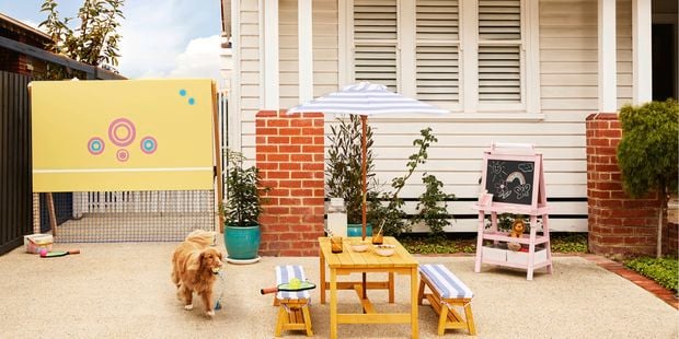 Dog carrying a toy walks across a paved front yard featuring a children’s tennis net, outdoor table with benches and umbrella, and a chalkboard easel in front of a weatherboard house.