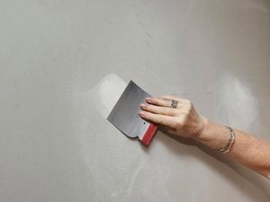 A hand prepares a white wall for painting using a scraper.