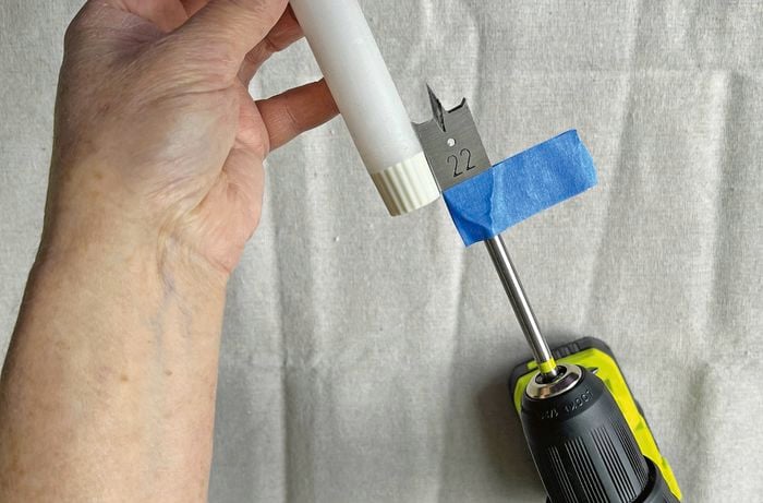 A hand holding a drill with blue painter’s tape wrapped around the spade bit on a drop sheet