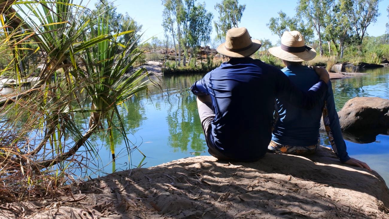 Rear view of couple sitting side by side on a large rock, looking out into a waterhole pool landscape