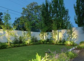 A well-lit landscaped garden with a green lawn, garden beds and a white fence
