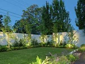 A well-lit landscaped garden with a green lawn, garden beds and a white fence