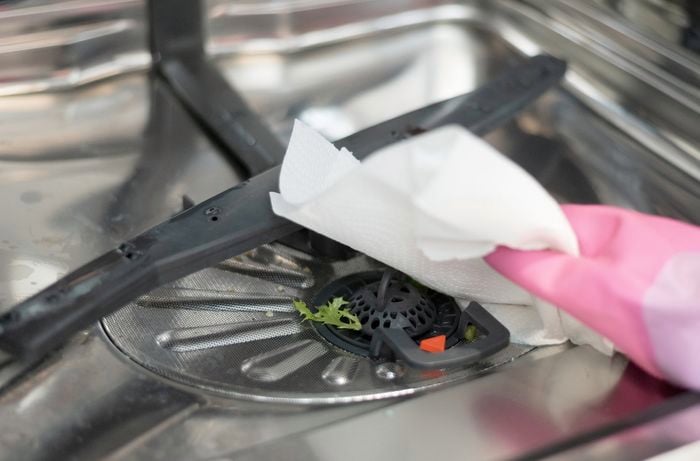 A person wearing pink gloves is cleaning the internal filter of the dishwasher using a paper towel and removing leftover food.