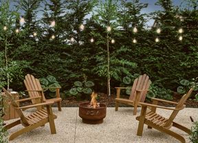 Outdoor fire pit with burning wood, surrounded by wooden chairs on a patio, under string lights and green trees.