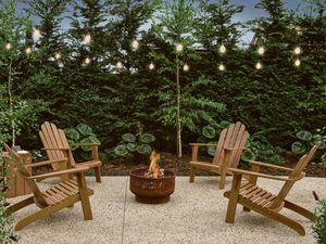 Outdoor fire pit with burning wood, surrounded by wooden chairs on a patio, under string lights and green trees.