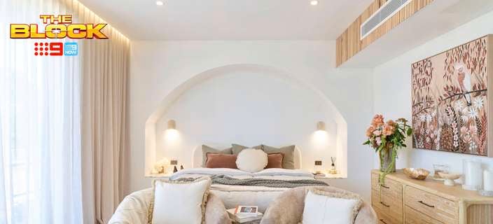 A furnished bedroom with a bed, two chairs, drawer's unit a painting and lights on the wall.