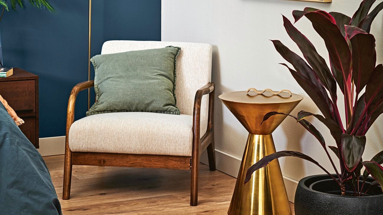 Bedroom with navy blue feature wall and white wall with painting hanging. White bedside chair and gold side table in between two, indoor pants.
