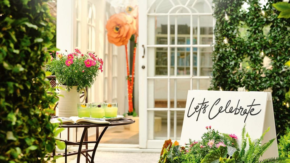 An outdoor patio with a bistro table, a planter of flowers, and a white door with a large paper flower decoration.