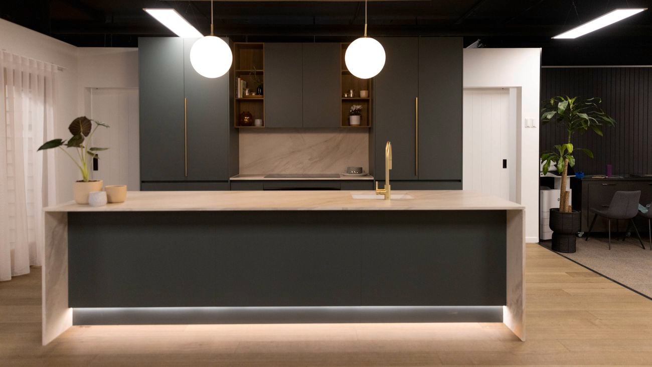 A wide-angle image of a Bunnings Building Solutions Studio kitchen set up with olive green paneling, gold tapware and handles, plus light-coloured splashback and benchtop