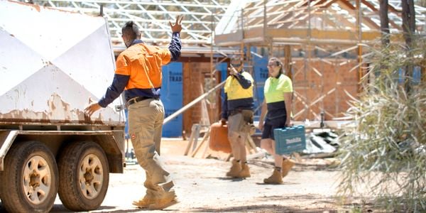 An image showing the back of a male tradie in orange hi-vis with one hand on the back of a work trailer and the other waving to two tradies – one male and one female – who are out of focus and carrying tools in boxes on a jobsite