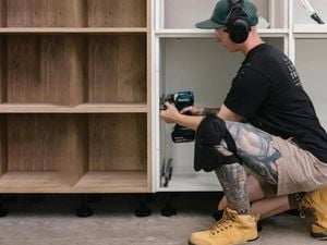 A tradesperson wearing a cap, earmuffs, and safety glasses kneels, using a power drill to install a white cabinet next to wooden shelving