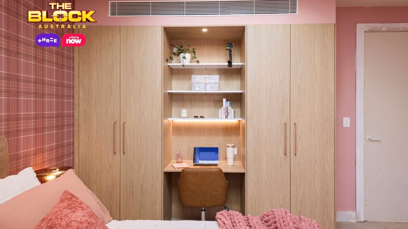 Pink bedroom with a check wallpapered wall and a double bed with pink linens. Opposite is a built in wardrobe with a desk in the middle.