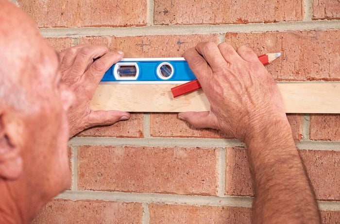 Person using spirit level and timber to mark out screw holes on brick wall.