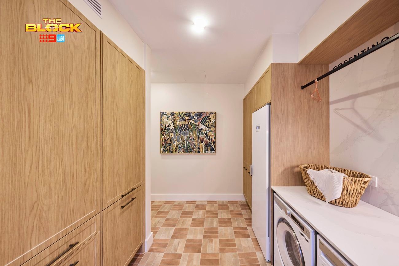 Laundry from The Block with timber cabinetry, tiled floor, washer and dryer, bench space, and wall art.