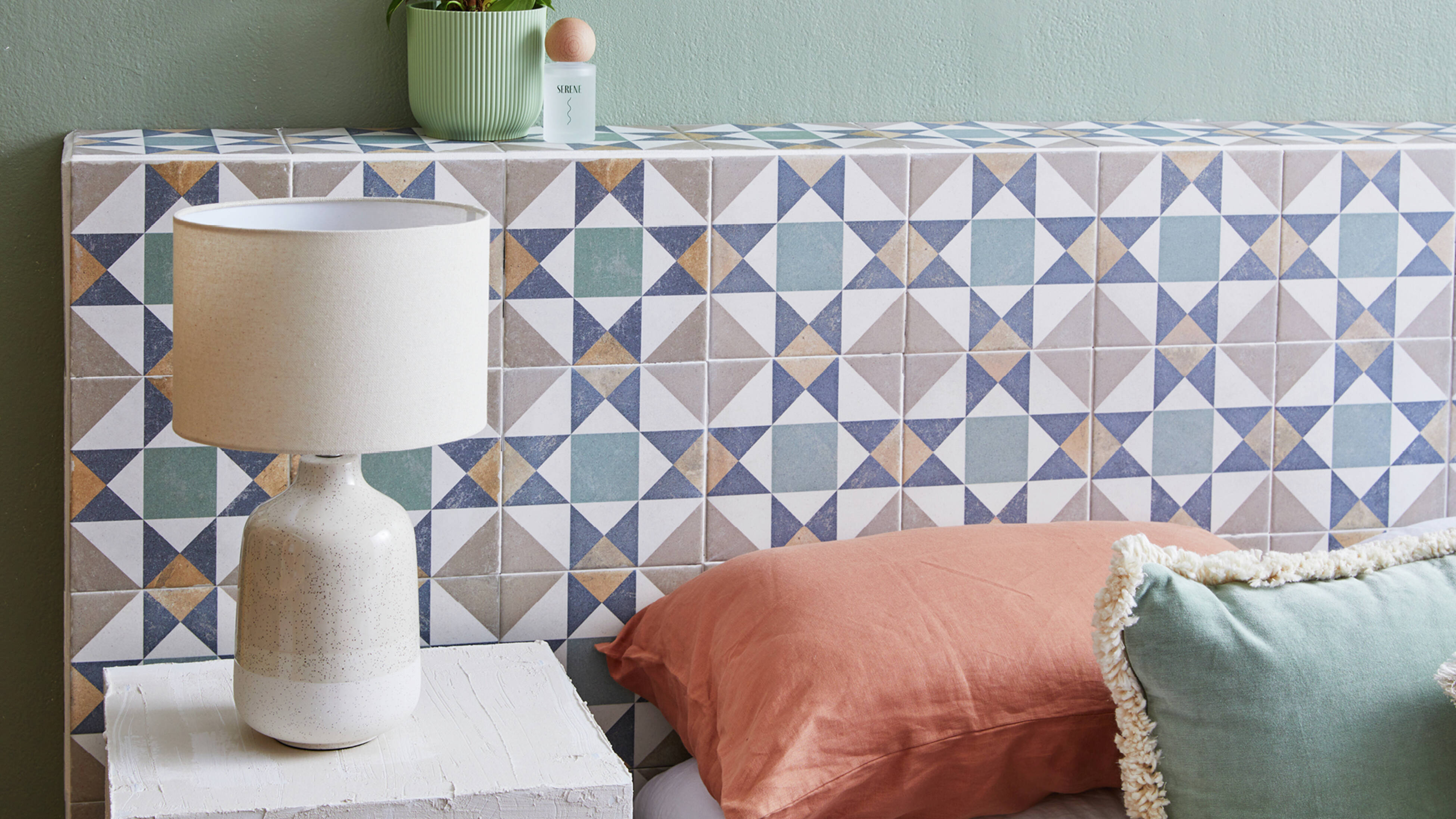 A bed with green and terracotta coloured pillows with a blue, grey, and white geometric tiled headboard. A white lamp on a white bedside table sits in front.] 