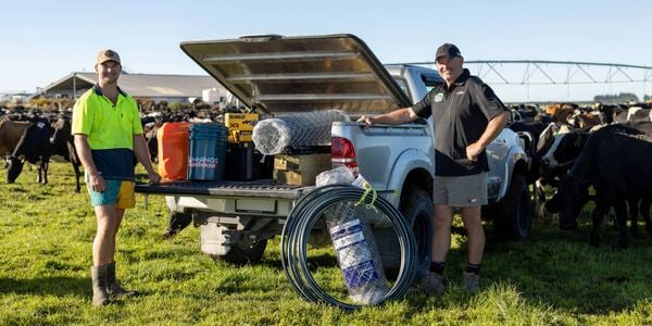 Two men standing beside an open ute on a farm, with cattle in the background; the vehicle is loaded with supplies including fencing wire, buckets, and tools.