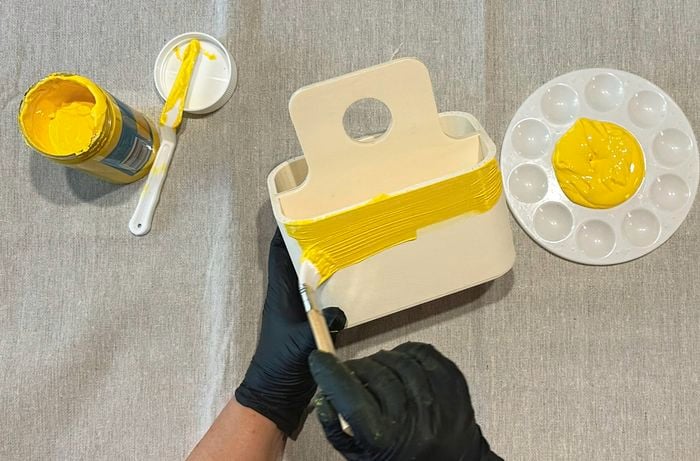 Hands wearing black gloves painting a wooden art caddy with bright yellow paint, with a jar of paint and palette beside it.