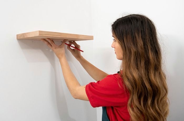Person holding a timber wall shelf against a wall and using a pencil to make a mark.