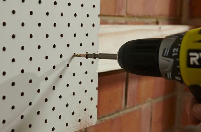 A pegboard being screwed to a mounted wooden length on a brick wall