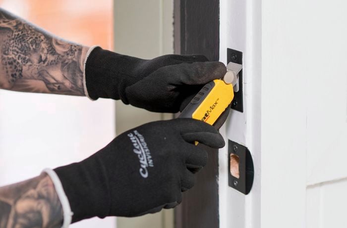 A person wearing gloves is using a utility knife to trace a strike plate on a door frame
