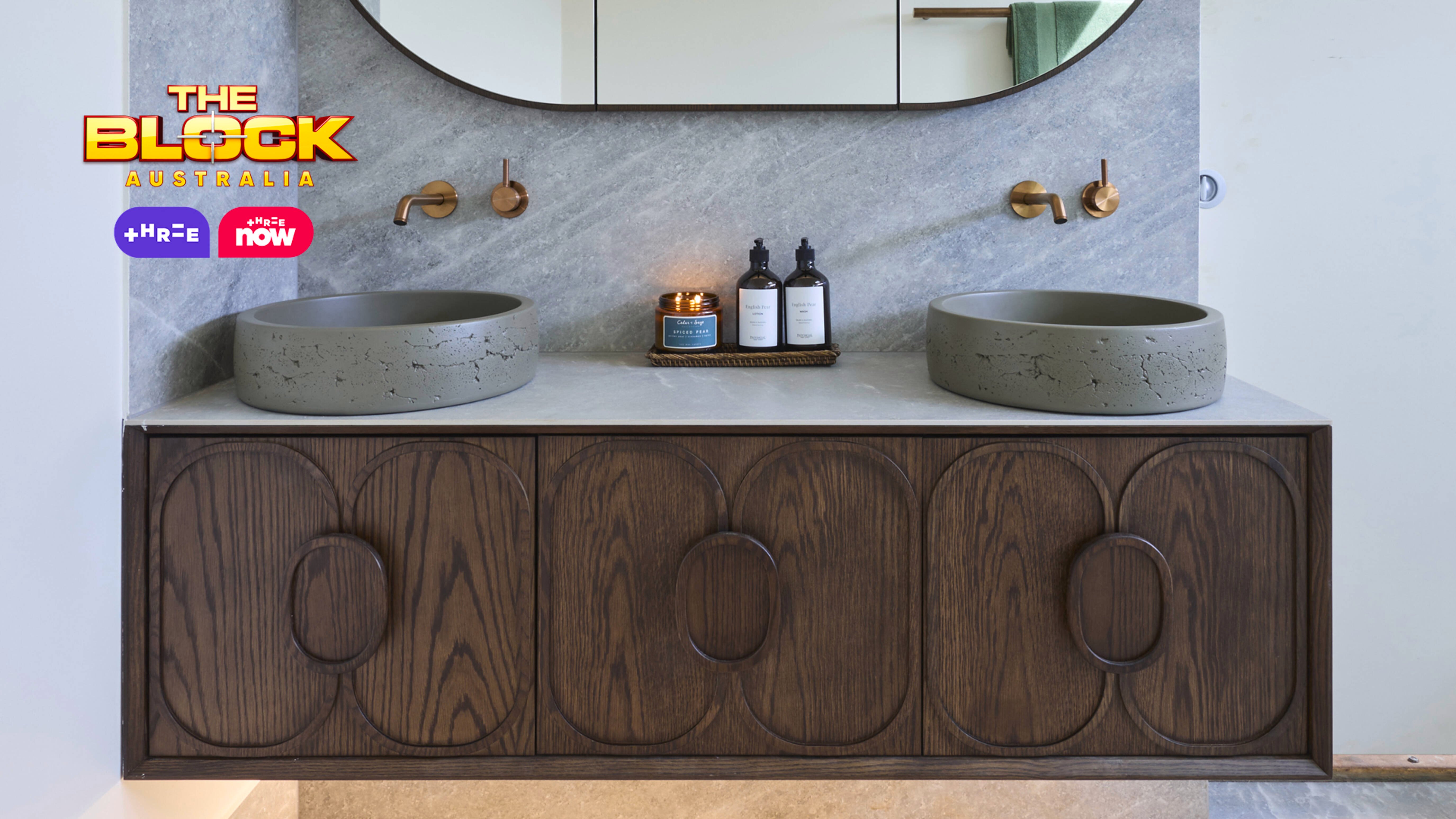 Modern bathroom with double stone sinks, brass wall-mounted faucets, oval mirror, marble countertop, dark wood vanity, and light stone tile floor. 