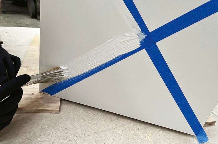 Gloved hand using a paintbrush to apply white paint along blue painter’s tape on a grey panel.