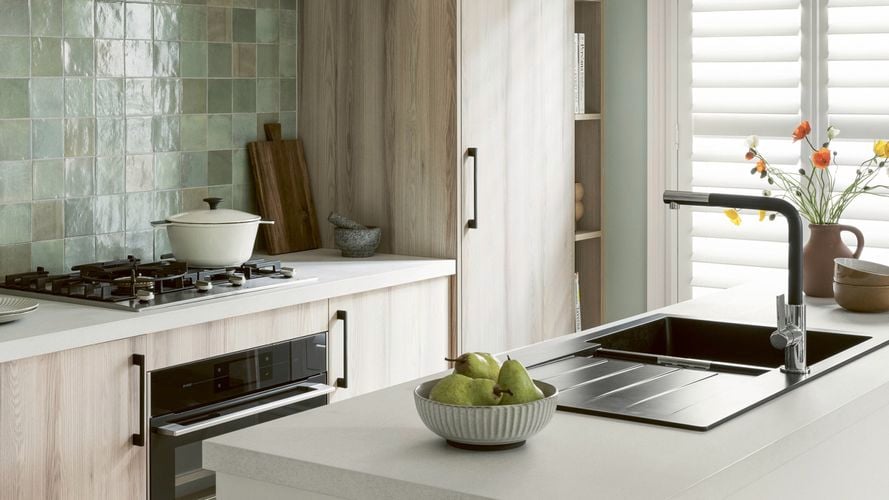 Modern kitchen with pale timber cabinets and white benchtops, a green tile splashback, a gas cooktop with a white pot, and a bowl of pears on the island