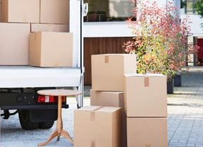  An open moving truck outside a modern house, filled with cardboard boxes and a white couch. Boxes and a small wooden table sit on the driveway.