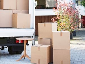  An open moving truck outside a modern house, filled with cardboard boxes and a white couch. Boxes and a small wooden table sit on the driveway.