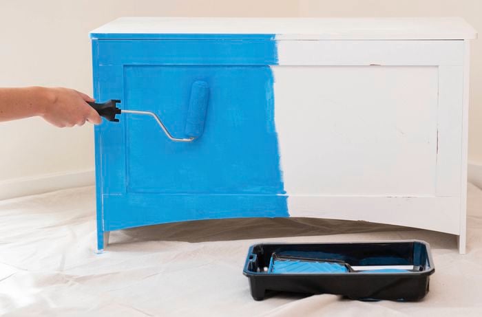 A person is painting a timber toy box with a bright blue base coat using a paint roller as part of a DIY games storage box makeover.