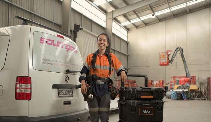 A lady in a trade uniform standing in front of a van in a garage with tools in it.