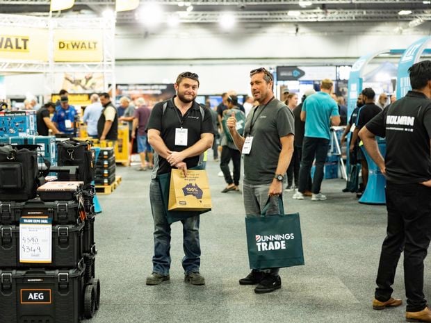 People at a trade expo talk near tool displays and branded stands in a large hall.