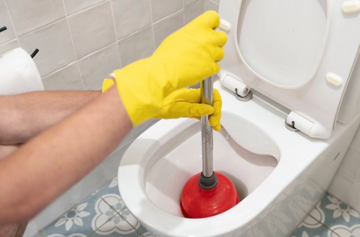 A person wearing yellow rubber gloves is using a plunger to unblock a toilet.