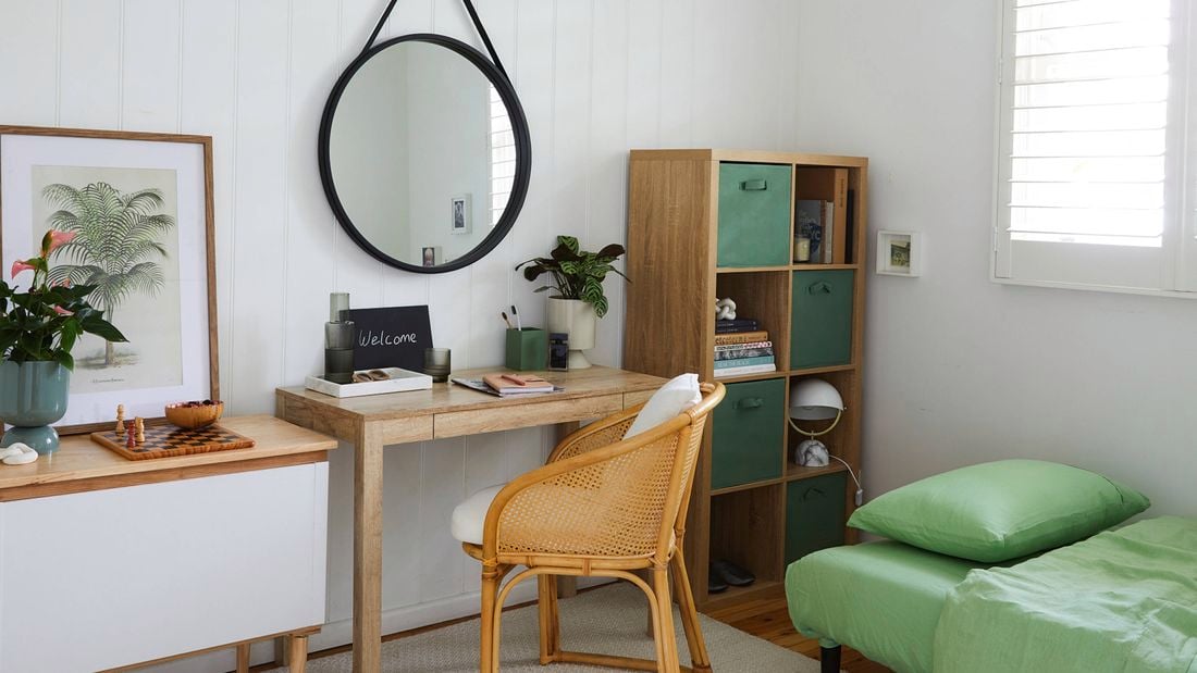 A bright guest room with white walls, a wooden desk and cane chair with a small plant and welcome chalkboard. To the right is a cube shelf with books and boxes, and a single bed with green bedding.