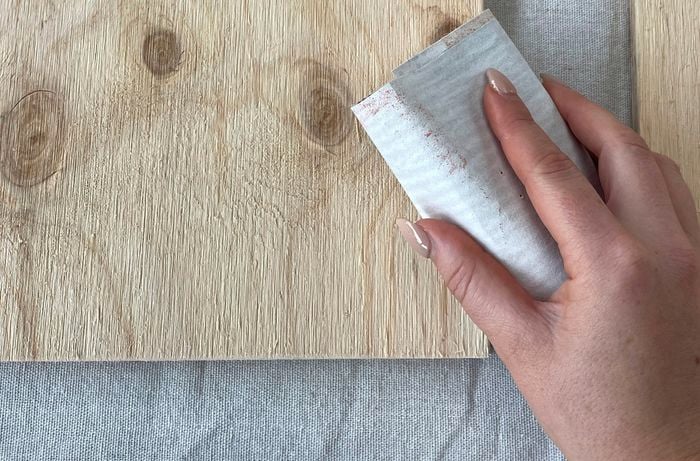 The pieces of plywood rest on the work surface while a hand is shown using sandpaper and a sanding block to smooth the cut edges